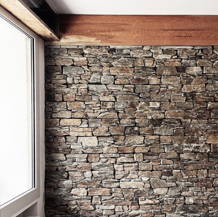 Mur intérieur en pierre naturelle fraîchement posé dans la salle de petit déjeuner du projet Le Jardin. Les pierres, aux teintes chaudes et variées, sont jointées avec soin. Un linteau en bois massif traverse le haut du mur, contrastant avec la texture minérale. À gauche, une grande baie vitrée laisse entrer la lumière naturelle, mettant en valeur la matière.